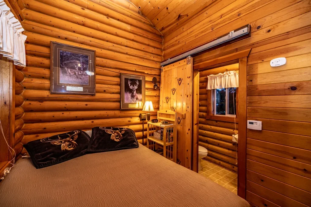 Small log cabin bedroom with a bed, wooden decor, framed deer art, and an open door to a bathroom.