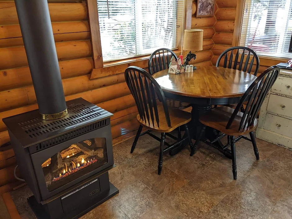A round wooden table with four chairs sits by windows in a log cabin, next to a lit wood stove.