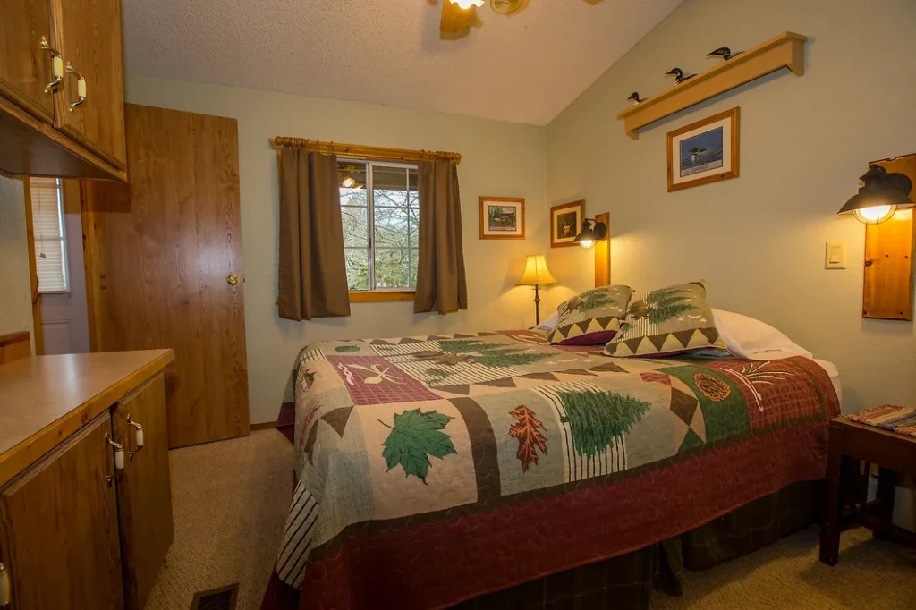 Small bedroom with a double bed, leaf-patterned quilt, wooden furniture, window with curtains, and wall art.