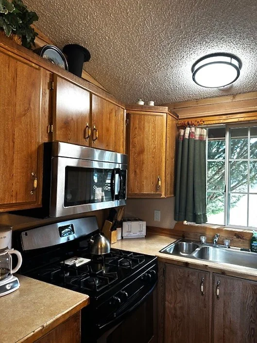 Small kitchen with wood cabinets, stainless steel microwave, gas stove, coffee maker, sink, and window with green curtains.