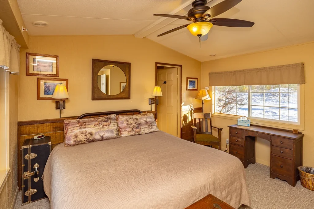 A cozy bedroom with a large bed, desk, chair, ceiling fan, wall lamps, and a window letting in natural light.