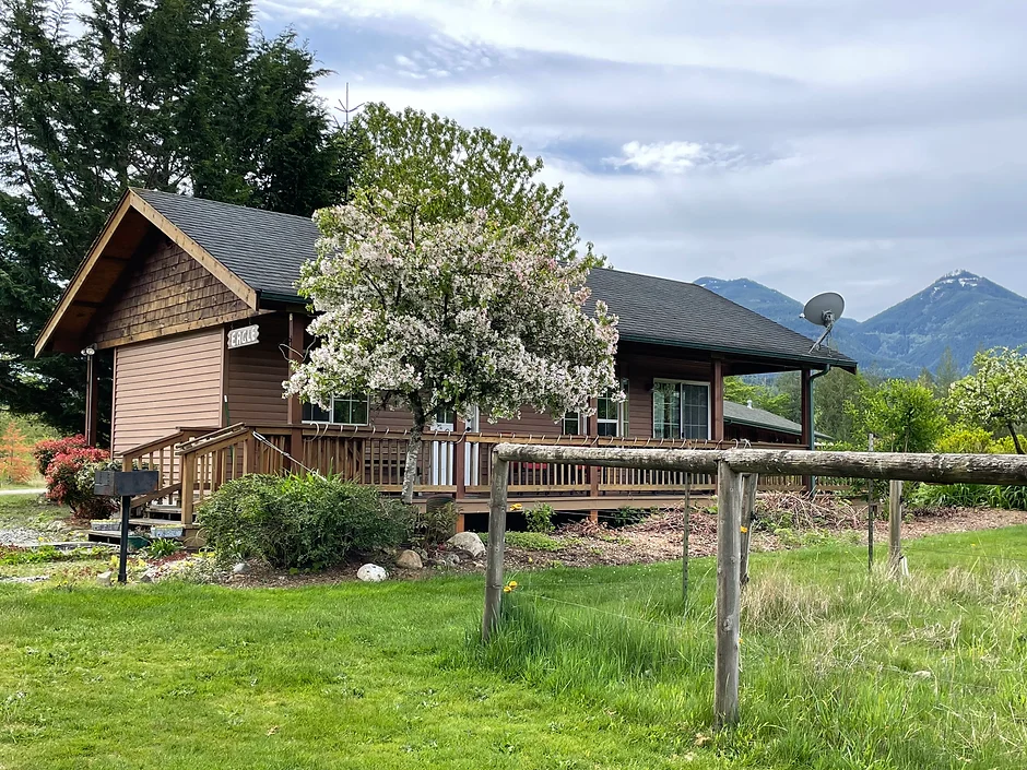 A brown house with a porch sits behind a blooming tree, with mountains and a cloudy sky in the background.