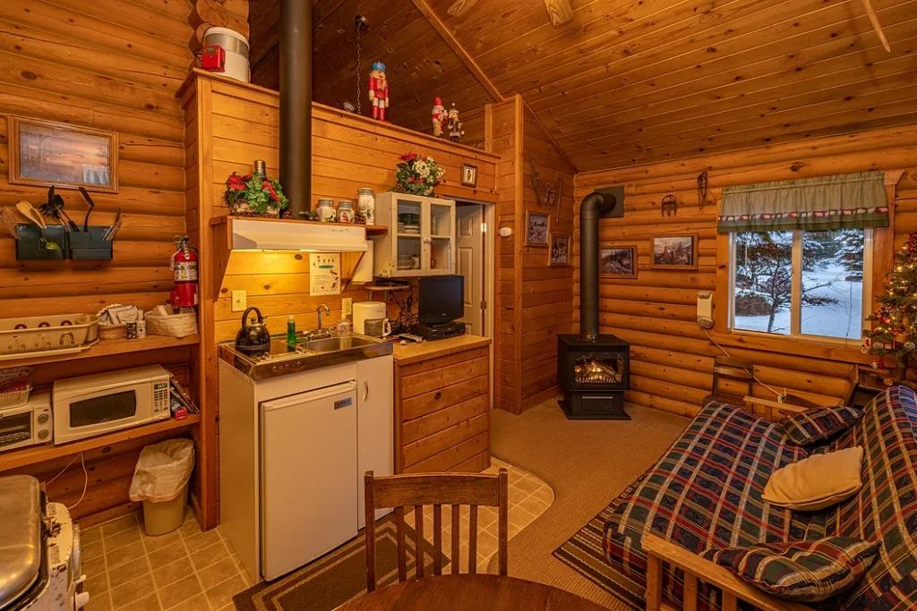 Cozy wooden cabin interior with a small kitchen, dining table, futon, wood stove, and Christmas decorations.
