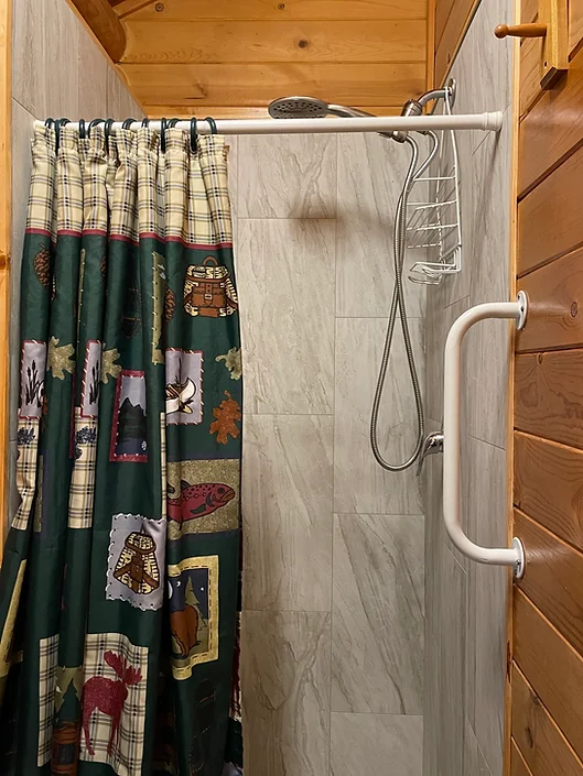 Small shower with a patterned curtain, wall-mounted showerhead, grab bar, and a metal shower caddy against tile walls.