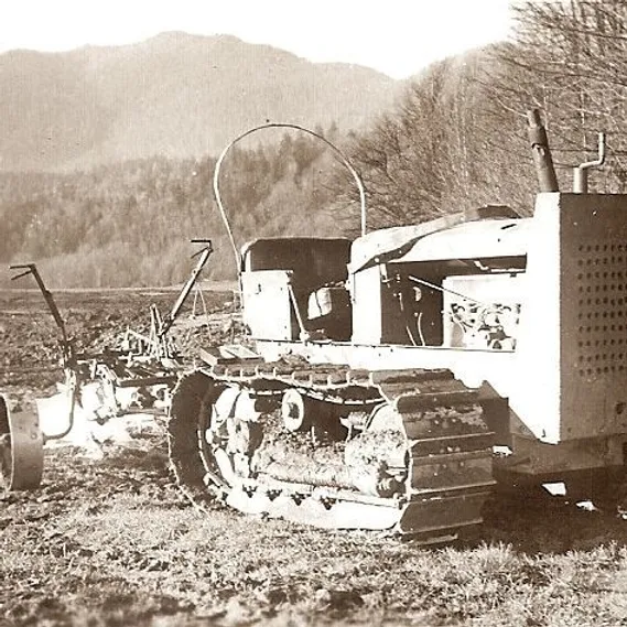 Vintage tracked tractor with plow attachment sits on grassy field with trees and mountains in the background.