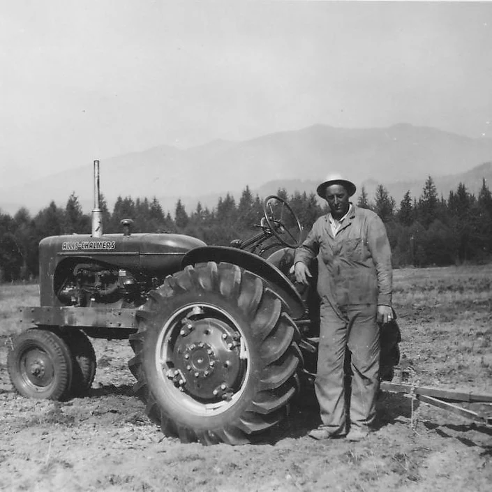 Man in overalls and hat stands beside an Allis-Chalmers tractor in a field with trees and mountains in the background.