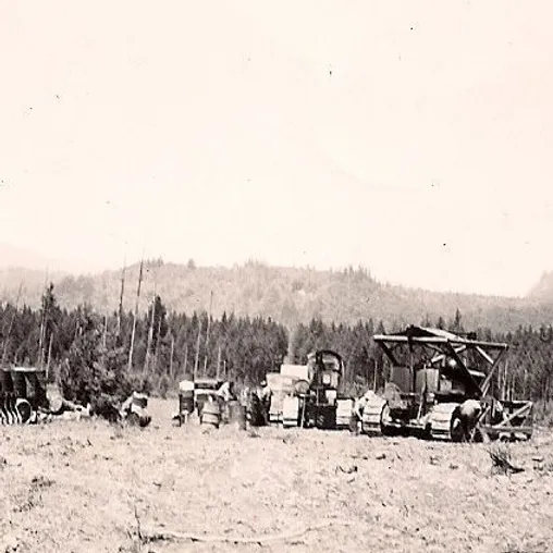 Vintage logging equipment and machinery are positioned in a cleared area with a forest and hills in the background.