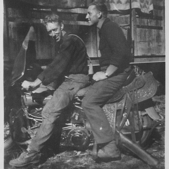 Two men sit side by side on a motorcycle, one at the handlebars, in front of a wooden structure.
