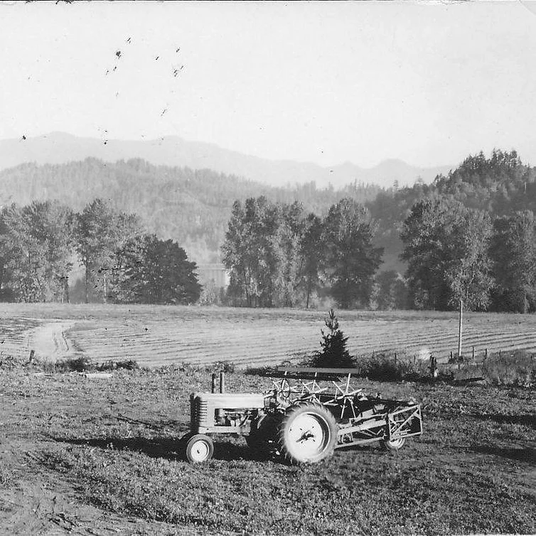 A tractor sits in a plowed field with trees and mountains in the background under a clear sky.